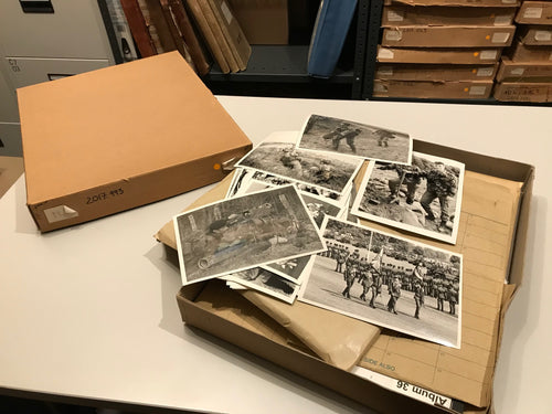 The image shows a shallow cardboard box open on a table on top are a series of black and white photographs, in the background are more boxes and albums on a shelf.