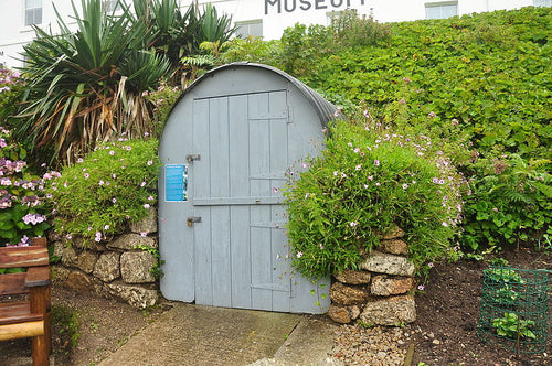 The image shows an Anderson shelter built at PK Porthcurno Museum of Global Communications. The shelter is painted grey with a corrugated metal roof, it is surrounded by a range of greenery.
