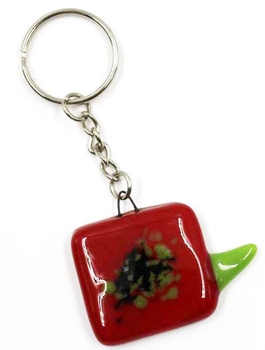 The image shows a square fused-glass poppy head of red glass with black and green centre and a green fused leaf element protruding off it. The poppy is attached to a silver-coloured metal keyring attachment.