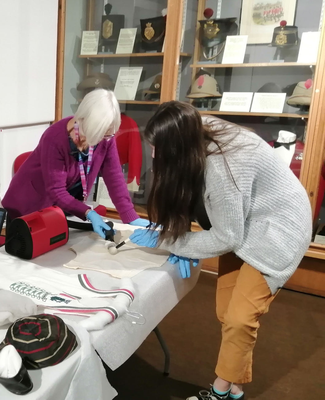 The image shows two female museum volunteers cleaning a vest in the museum gallery using a small red and black museum vacuum cleaner.