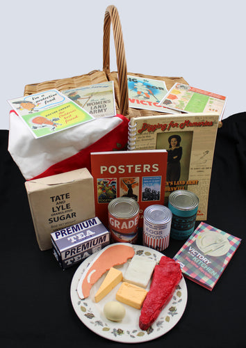 The image shows a wicker basket in from of which are books relating to the Second World War as well as food tins and food packaging as well as replica food on a ceramic plate. On top of the basket is an apron and pictures of posters from the Second World War.